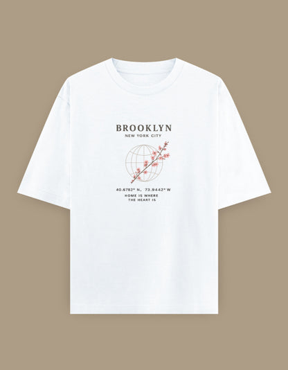 White oversized t-shirt featuring minimalist Brooklyn New York typography with cherry blossom artwork and globe-inspired line art printed on the front.